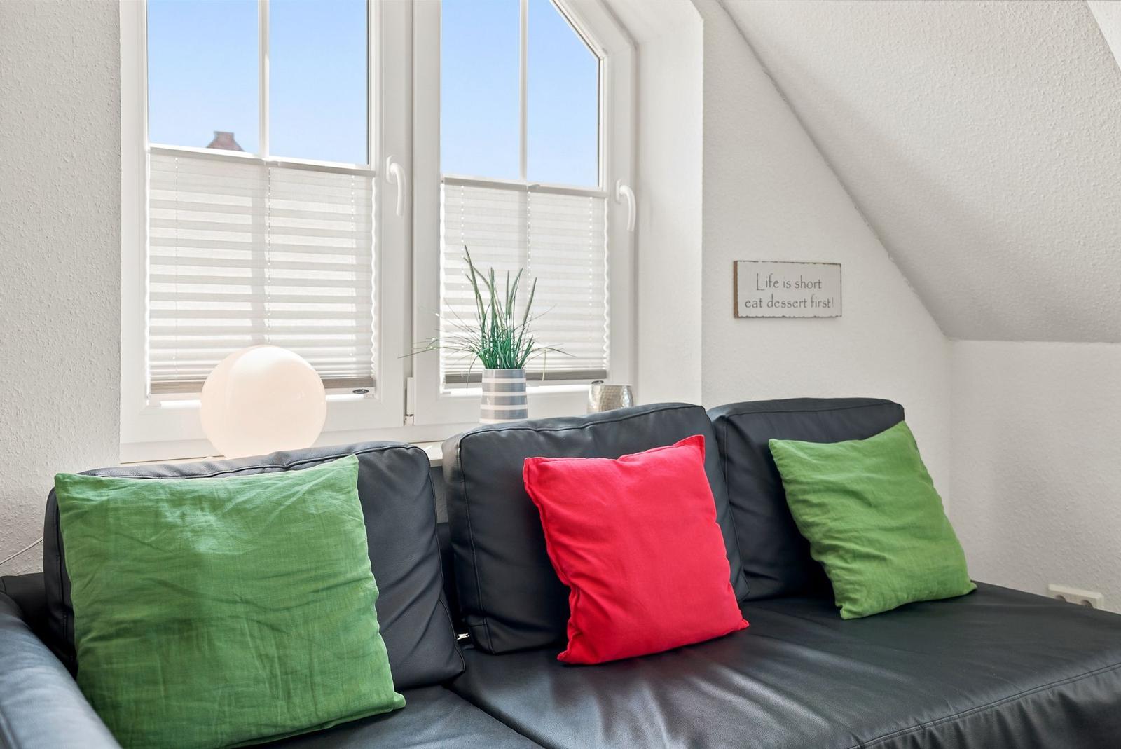 Schwarzes Sofa mit grünen und roten Kissen vor einem Fenster mit Jalousien.