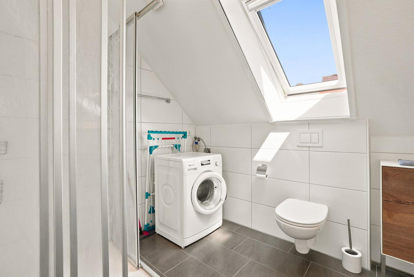 Dusche mit Waschmaschine, WC und Dachfenster. Weiße Fliesen, grauer Boden.