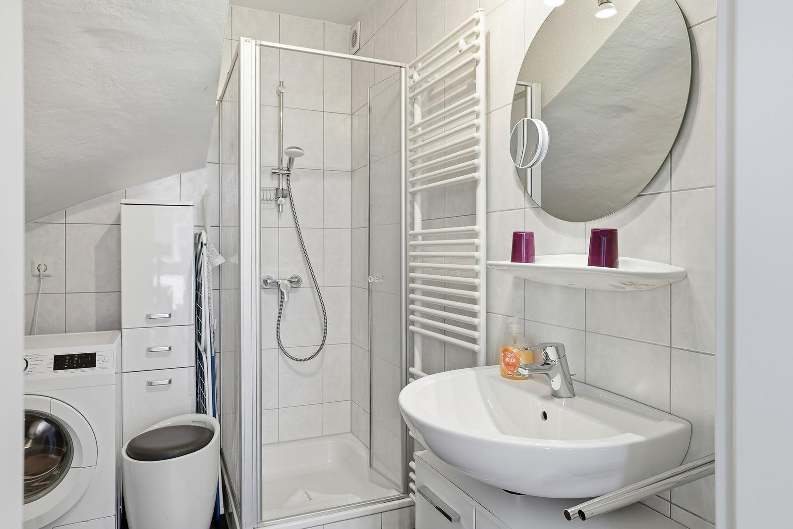 Badezimmer mit Dusche, Waschbecken und Wärmeelement. Weiße Fliesen und Spiegelschrank.