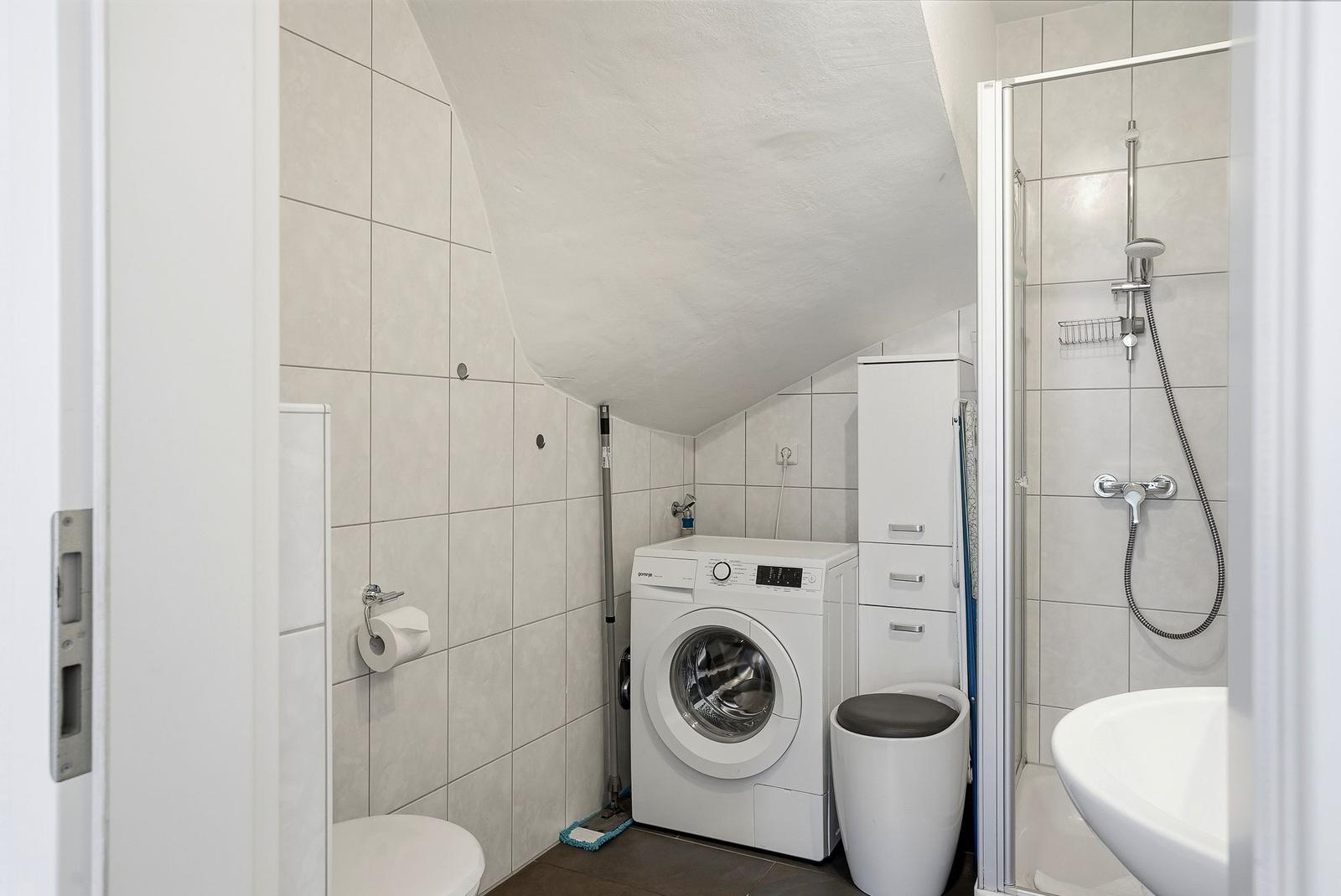 Badezimmer mit Waschmaschine, Dusche und Toilette unter schrägem Dach.