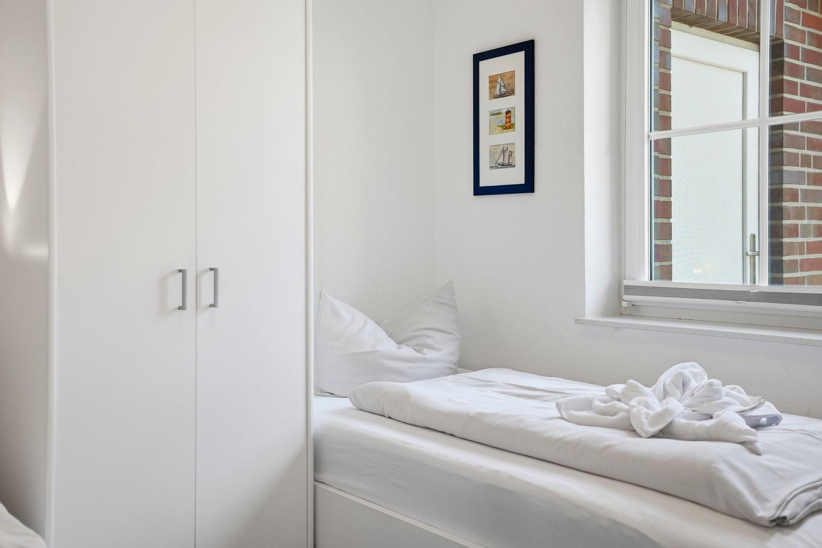 Schlafzimmer mit weißem Bett, Kleiderschrank und Fenster mit Blick auf Backsteinmauer.