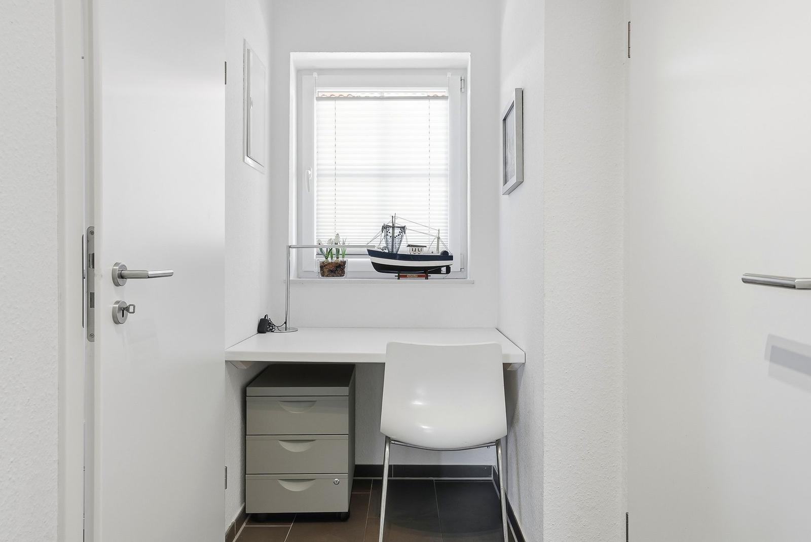 Arbeitsplatz mit Schreibtisch, Stuhl und Fenster. Auf dem Fensterbrett steht ein Schiffmodell.