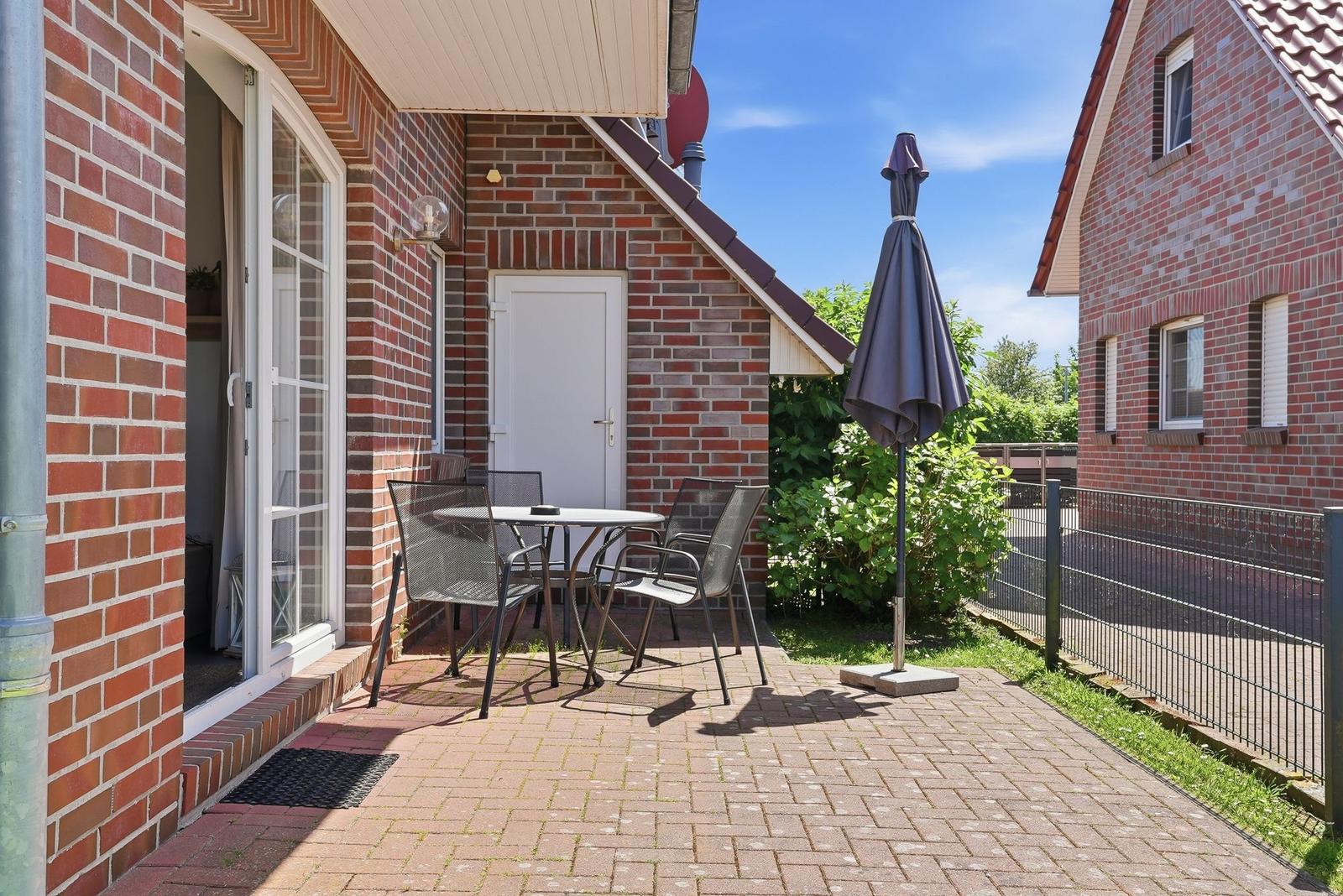 Terrasse mit Tisch, Stühlen und geschlossenem Sonnenschirm neben rotem Backsteinhaus.