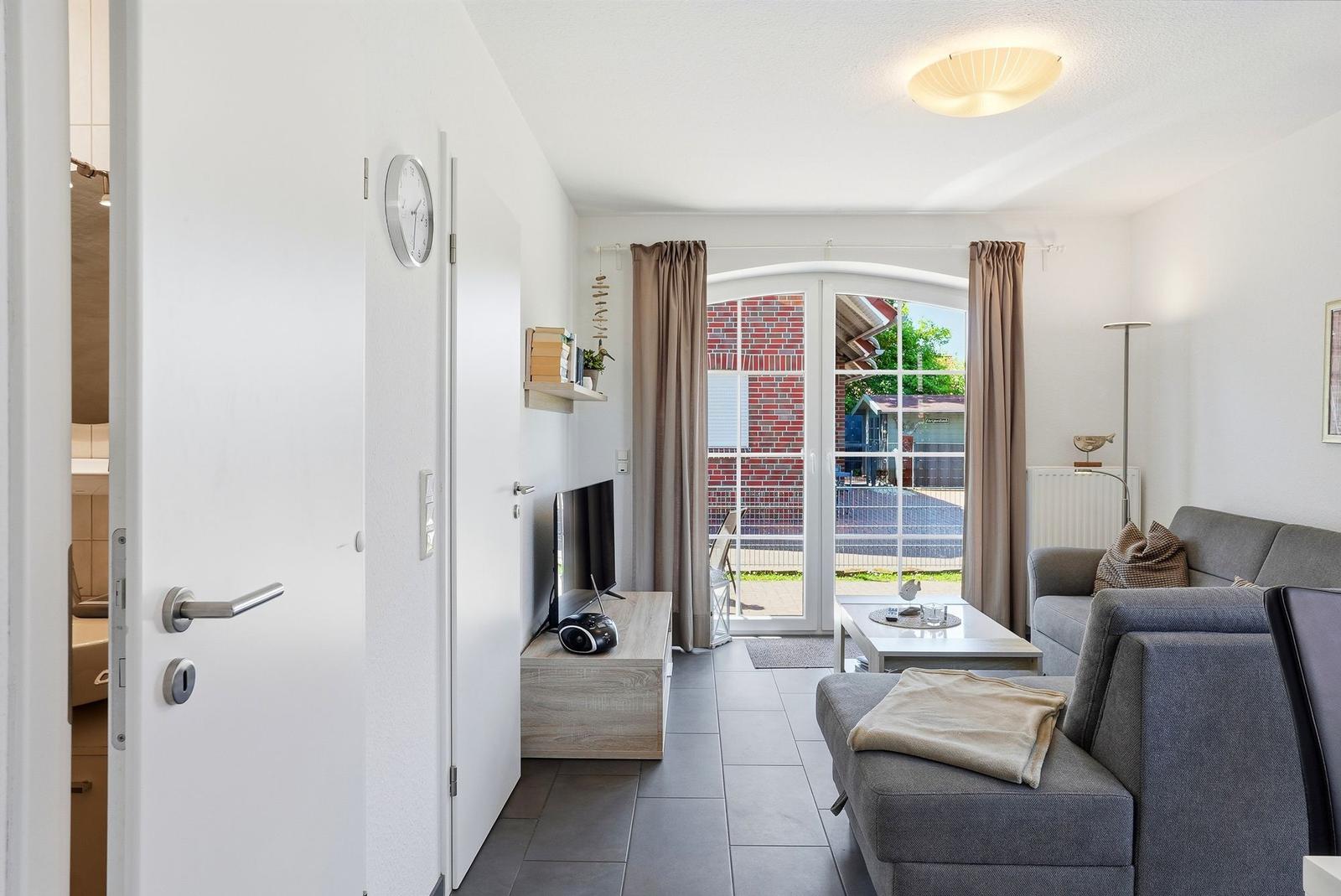 Wohnzimmer mit Sofa, Couchtisch und TV. Durchgang zur Küche. Fenster mit Blick auf Außenbereich.