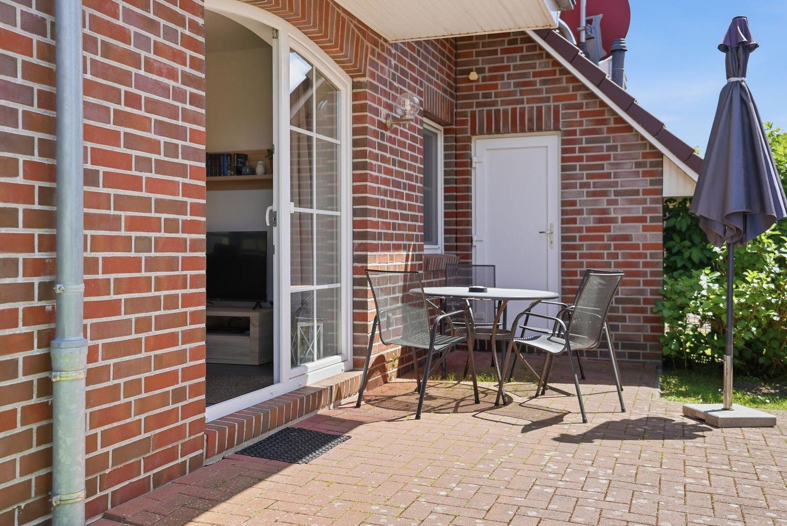 Terrasse mit Tisch, Stühlen und Sonnenschirm. Durch Fenster ist Wohnzimmer sichtbar.