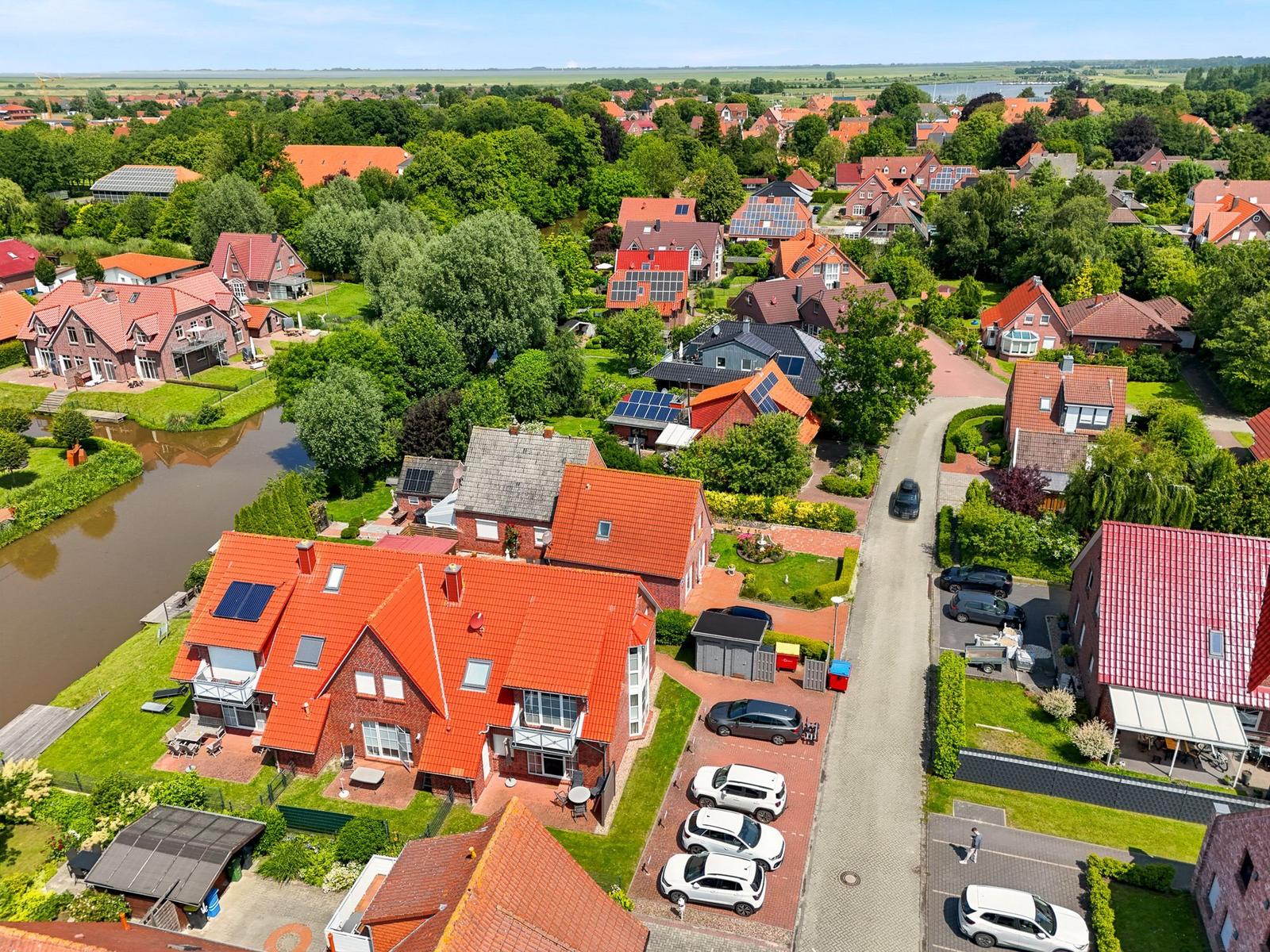 Haus mit rotem Dach und Garten neben Wasser. Mehrere Autos parken vor dem Haus.