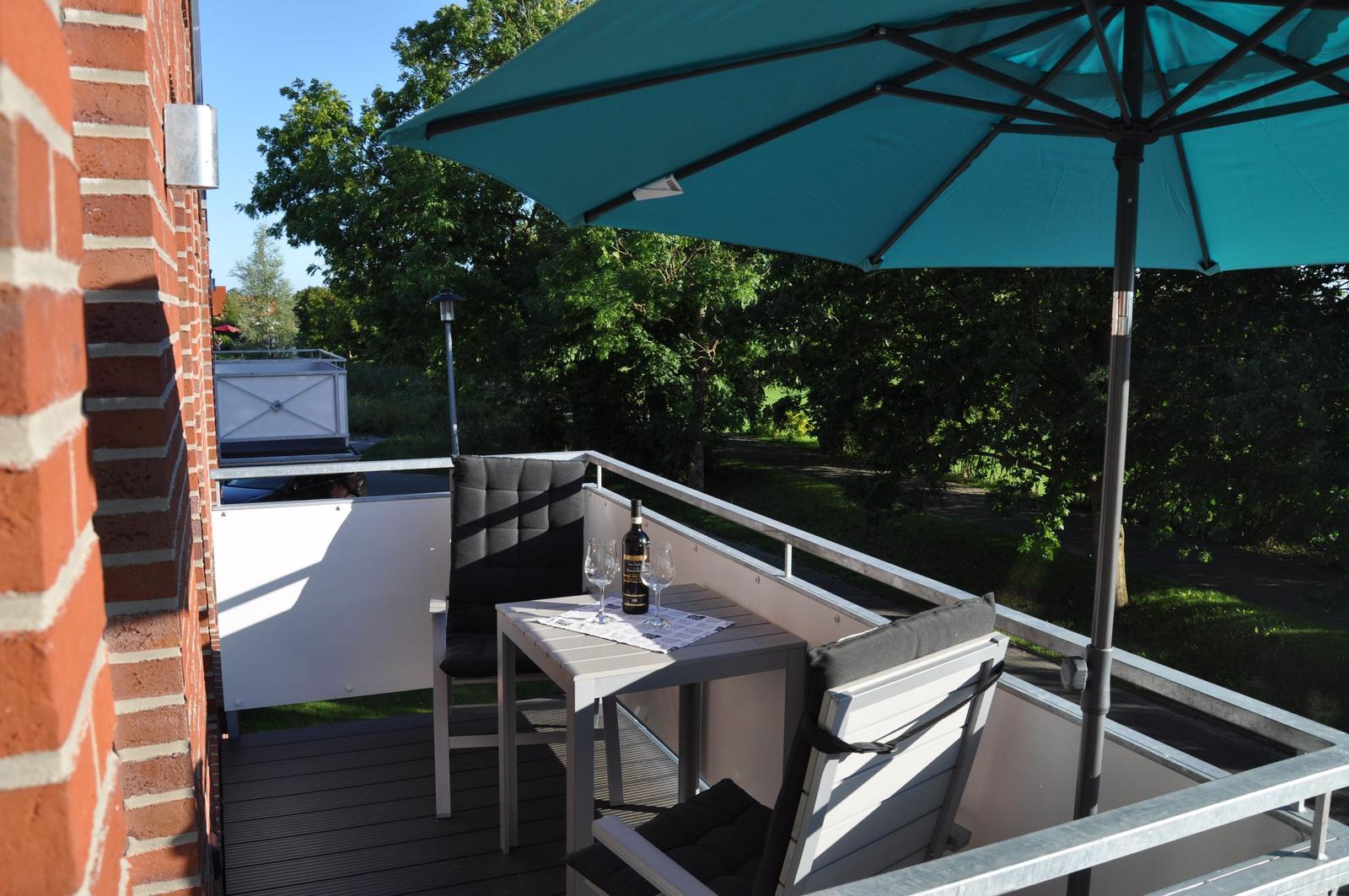 Terrasse mit Tisch, zwei Stühlen und einem blauen Sonnenschirm. Blick auf grüne Bäume.