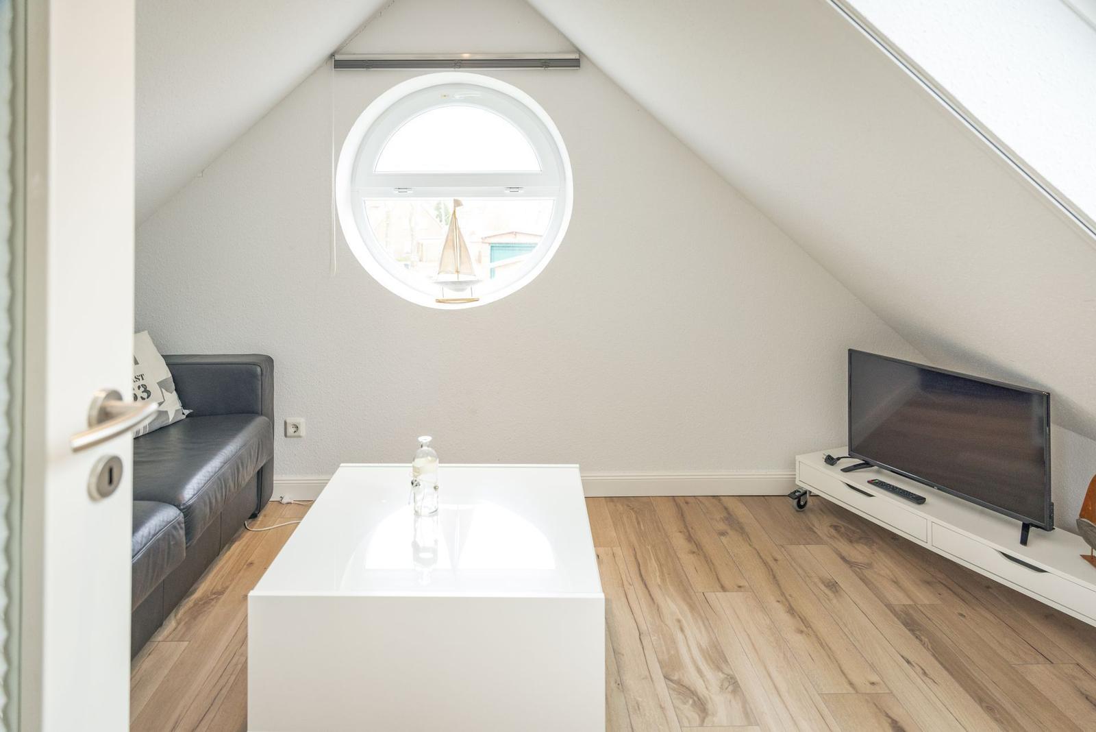 Wohnraum mit runden Fenster, Sofa, TV und weißem Tisch auf Holzboden.