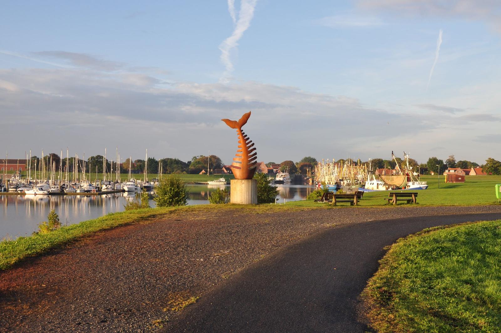 Weg führt an einer Whalskulptur vorbei zum Hafen mit Booten und grünen Wiesen.