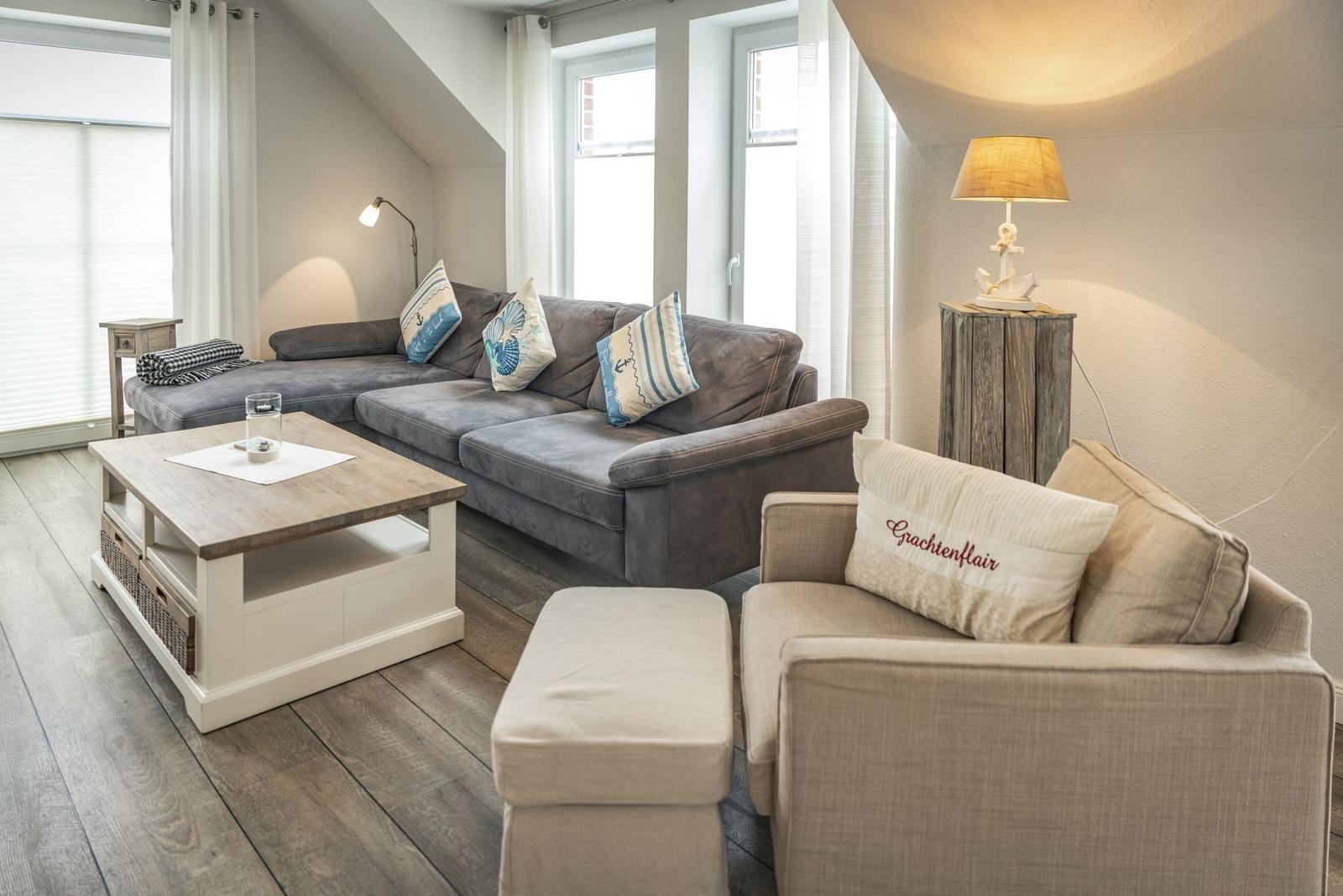 Wohnzimmer mit grauem Sofa, Beistelltisch und Sessel. Holzfußboden und Fenster mit Vorhängen.