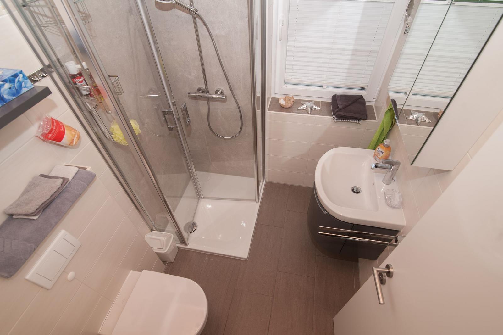 Badezimmer mit Dusche, Waschbecken und Toilette. Fenster mit Jalousie.