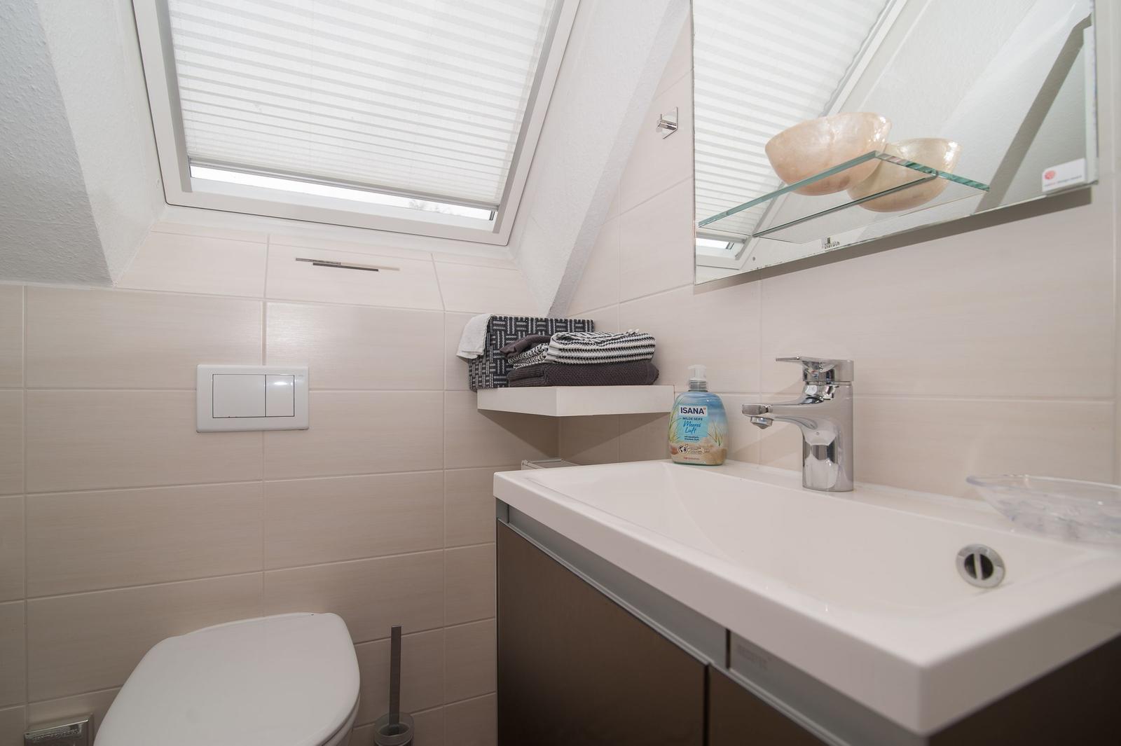 Badezimmer mit Duschwanne, Spiegel und Fenster. Toilette und Handtuchregal sind sichtbar.