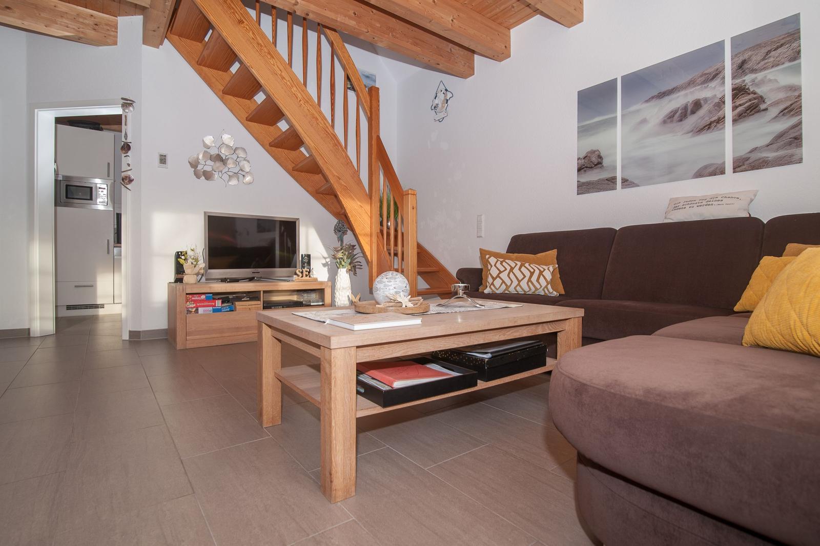 Wohnzimmer mit braunem Sofa, Holztisch und Holztreppe. Küche im Hintergrund.