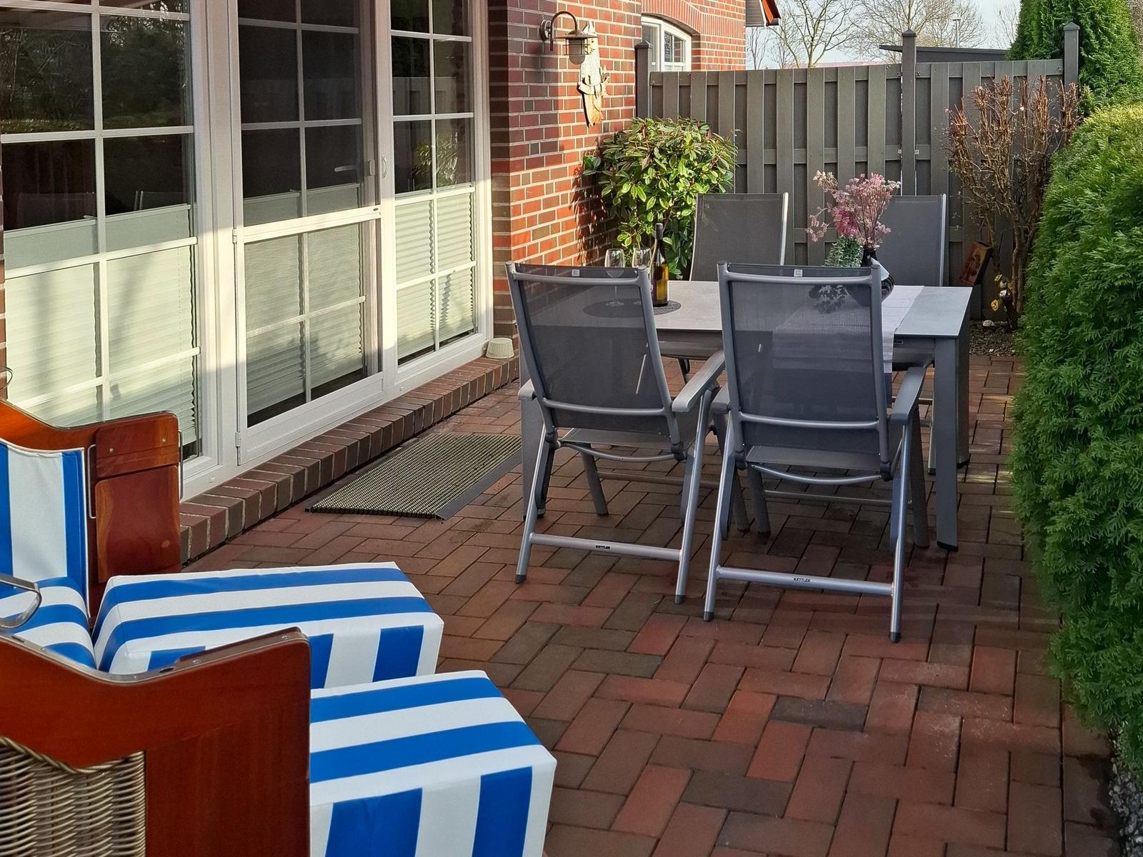 Terrasse mit Tisch, Stühlen und blau-weißem Liegestuhl. Hinterhof mit Zaun und Pflanzen.