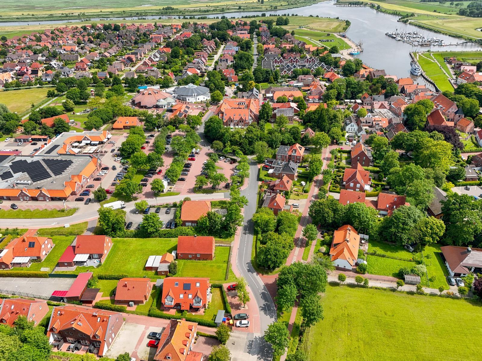 Luftbild eines Dorfes mit roten Dächern, Grünflächen und einem Fluss mit Booten.