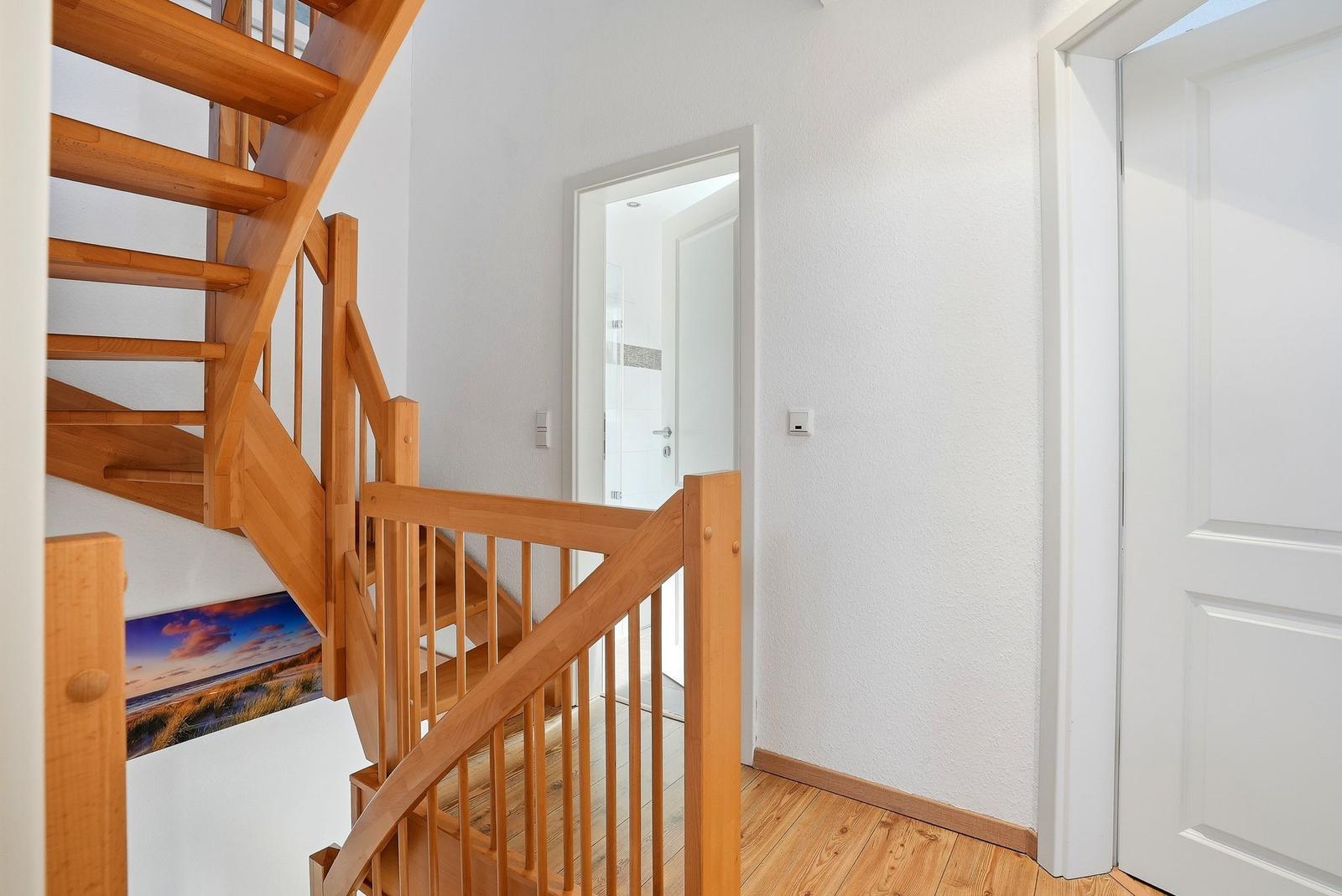 Holztreppe mit Geländer führt zu den oberen Etagen. Weiße Wände und Tür. Wandbild mit Landschaft.