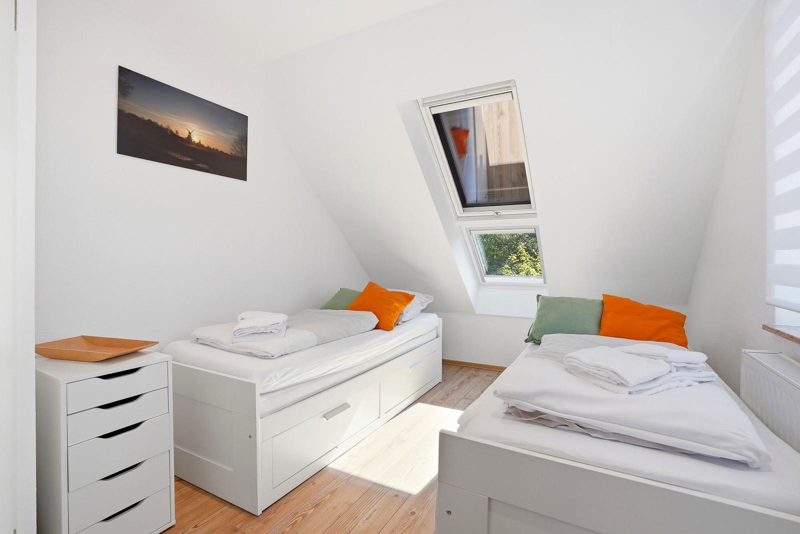 Zwei Betten mit weißen Bettwäschen und orangefarbenen Kissen. Dachfenster und Holzboden.