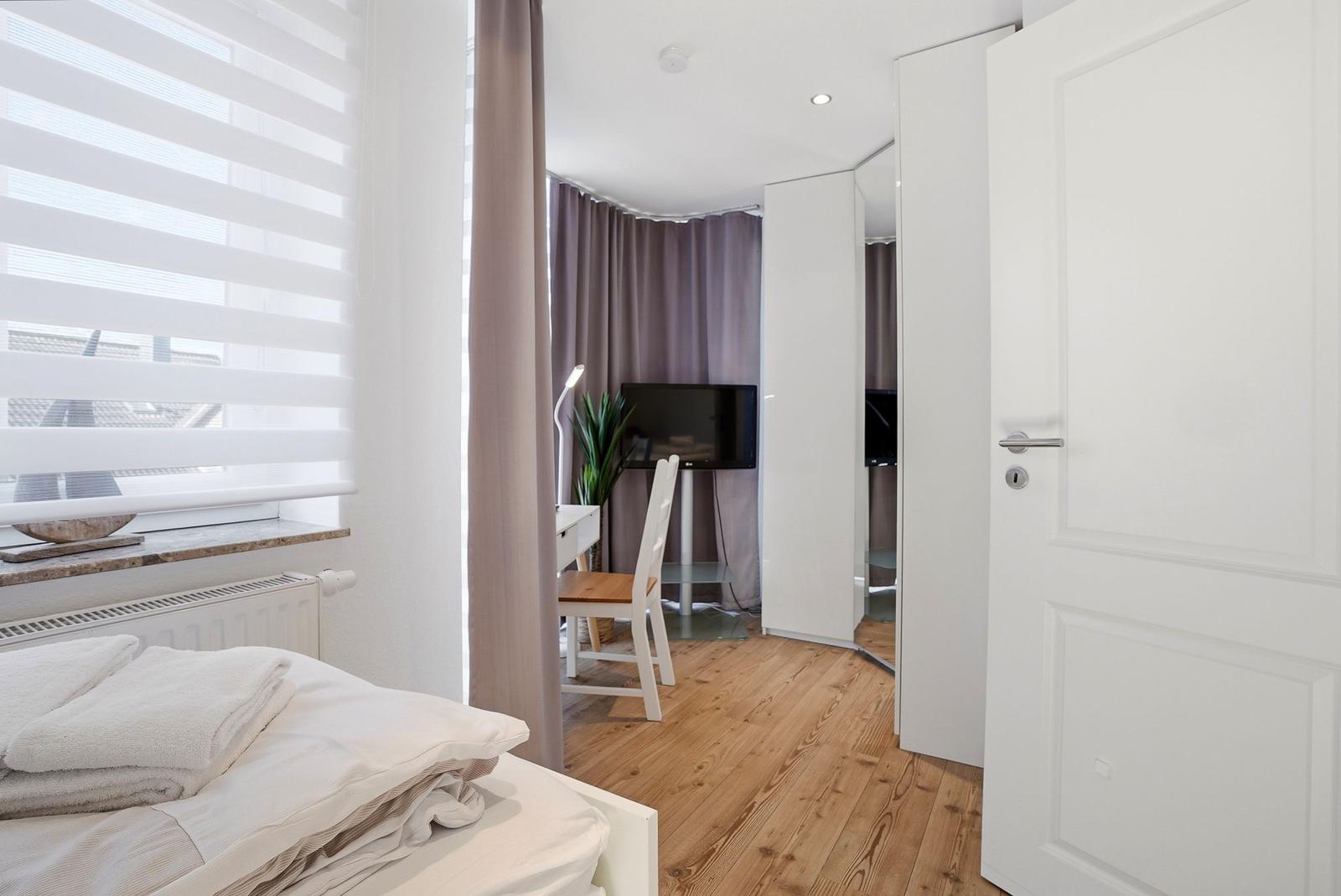 Schlafzimmer mit Bett, Fenster und Blick in Arbeitsbereich mit TV und Schreibtisch.