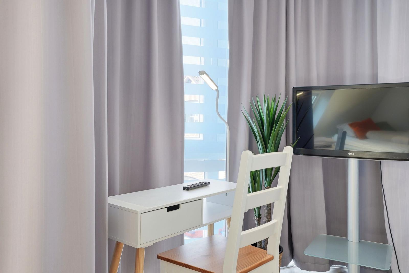 Schreibtisch mit Stuhl, Fernseher und Pflanze vor Fenster mit Vorhängen.