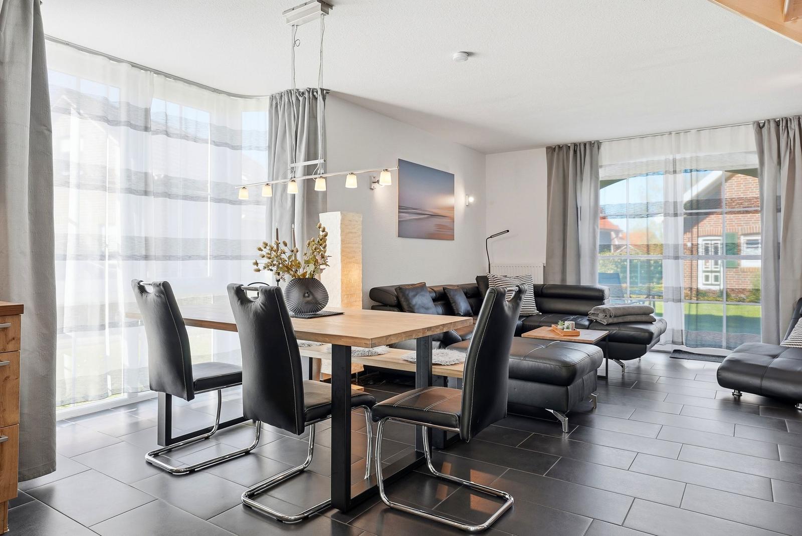 Essbereich mit Tisch und Stühlen, Wohnbereich mit Sofa und Couchtisch, große Fenster mit Gardinen.