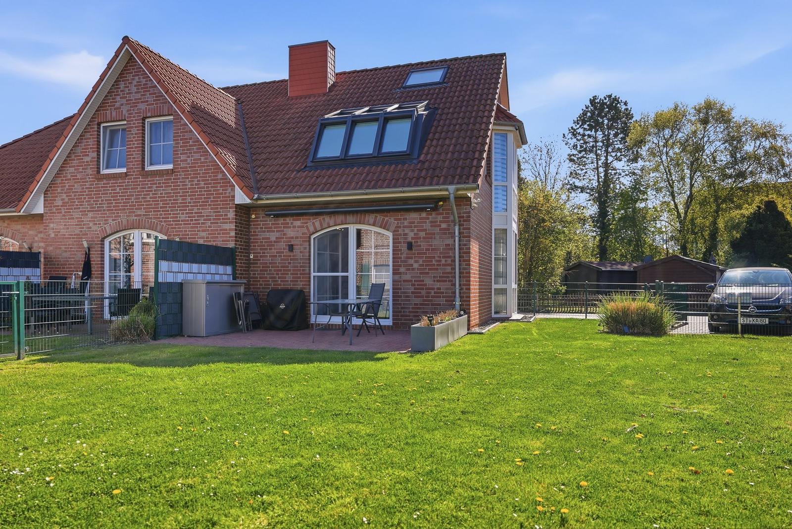 Rotes Ziegelhaus mit Terrasse und grünem Garten. Hinterhof mit Zaun und Auto.