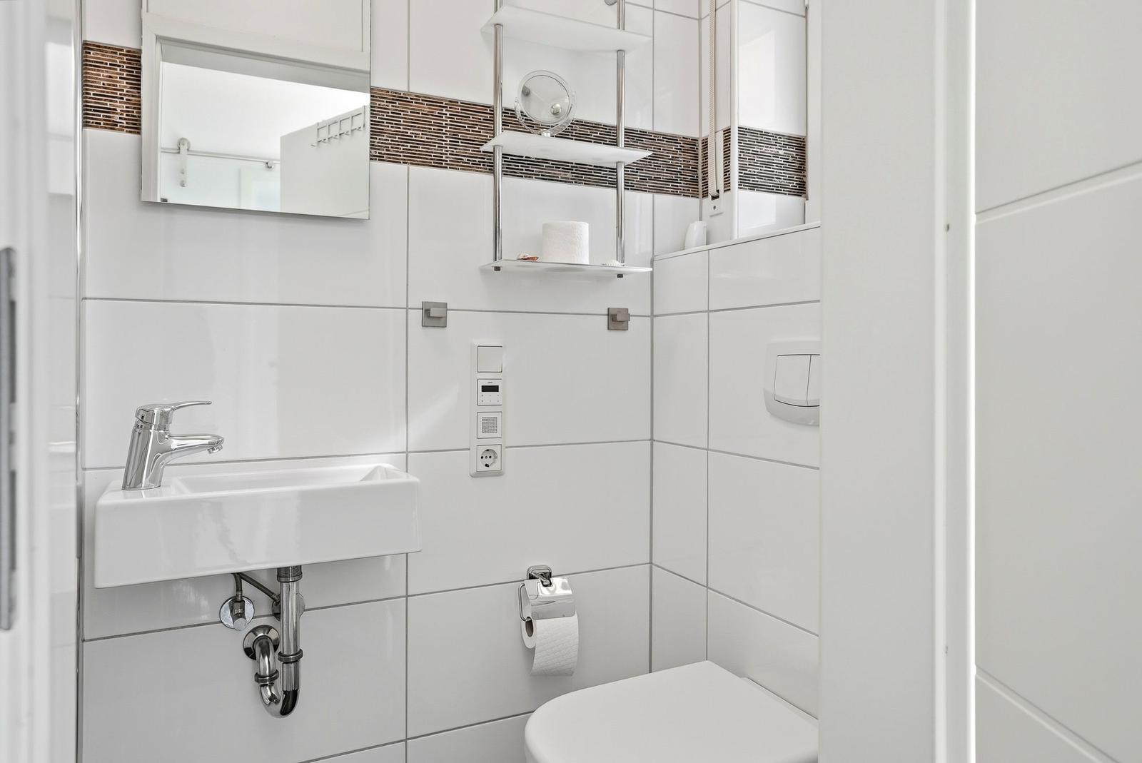 Badezimmer mit Waschbecken, Spiegel und Toilette. Weiße Fliesen und Metallregal.