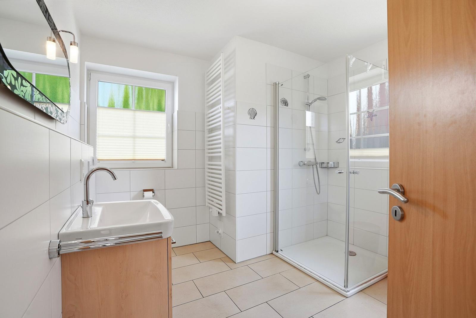 Badezimmer mit Dusche, Waschbecken und Fenster. Weiße Fliesen und Holzakzente.