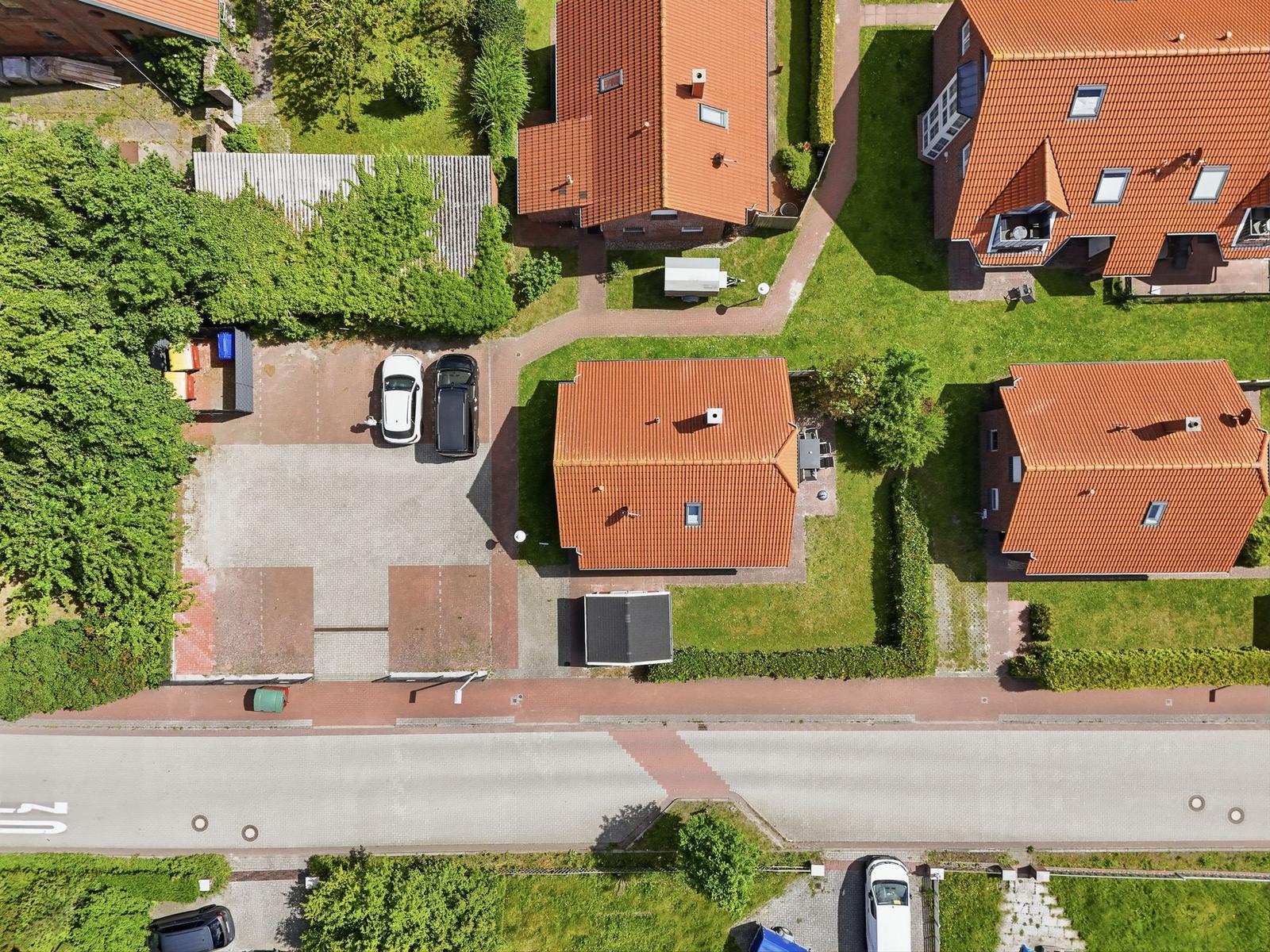 Luftbild von Wohnhäusern mit roten Dächern und grünen Gärten. Parkplätze und Straßen sind sichtbar.