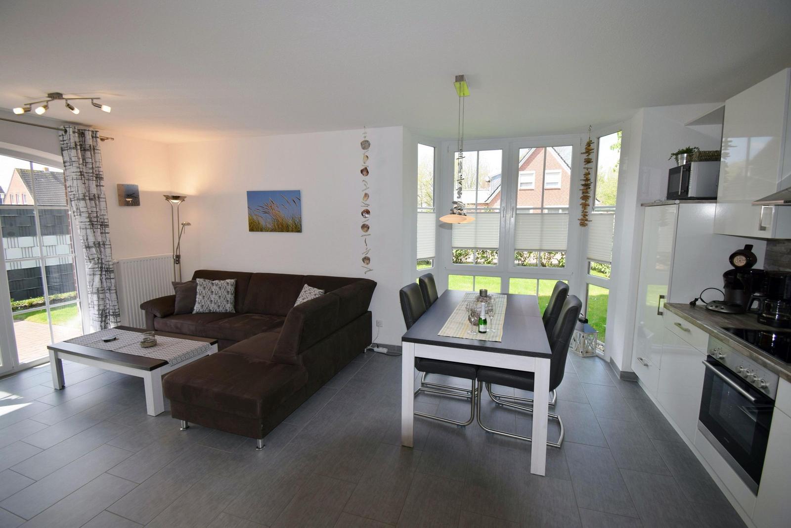 Wohn- und Essbereich mit Sofa, Esstisch und Küche. Große Fenster mit Blick auf Garten.