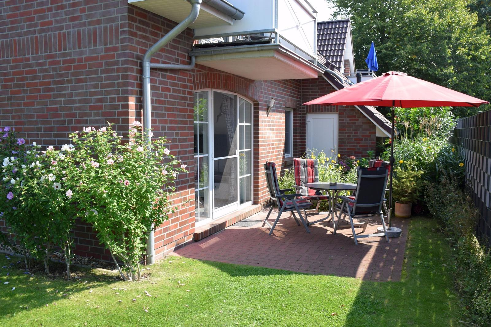 Roter Sonnenschirm über Tisch und Stühlen auf Terrasse neben Haus mit Blumenbeete.