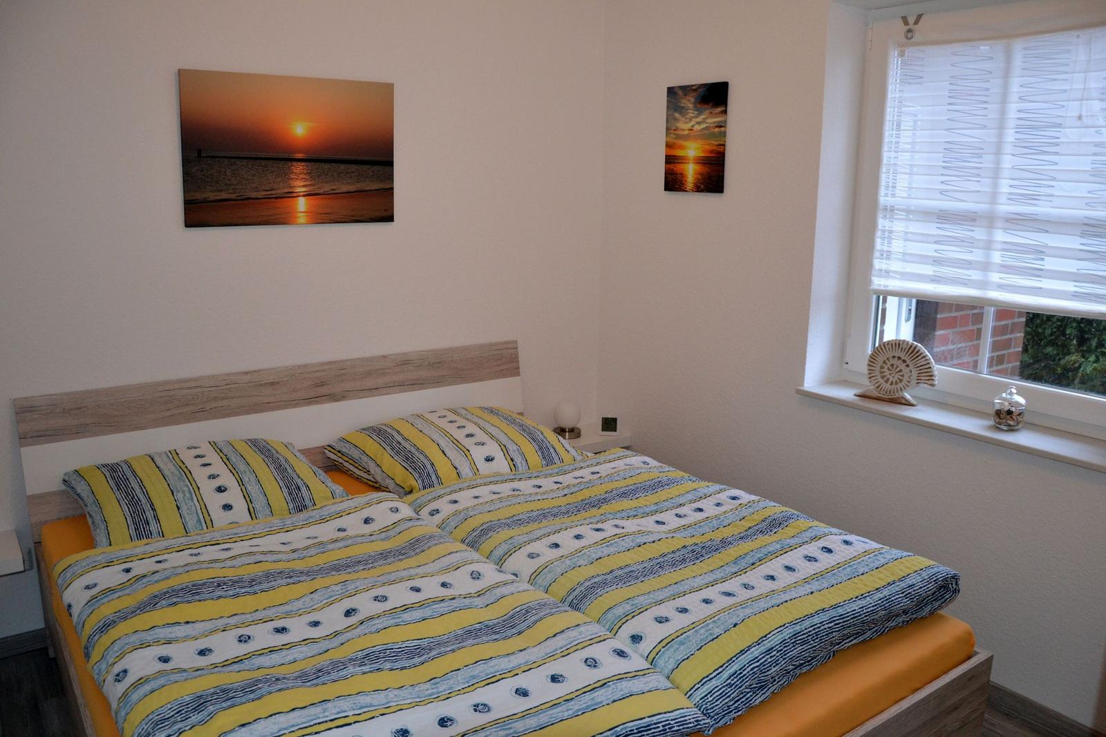 Schlafzimmer mit Doppelbett, Wandbildern und Fenster mit Jalousie.