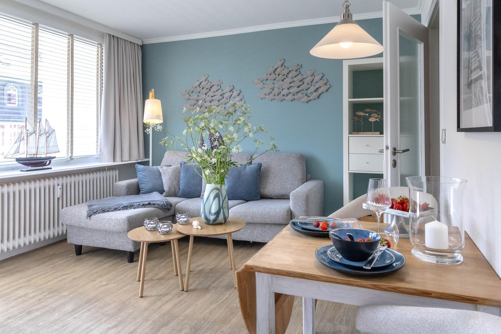 Wohn- und Essbereich mit Sofa, Tisch und Fenster. Wanddekoration und Blumen sind sichtbar.