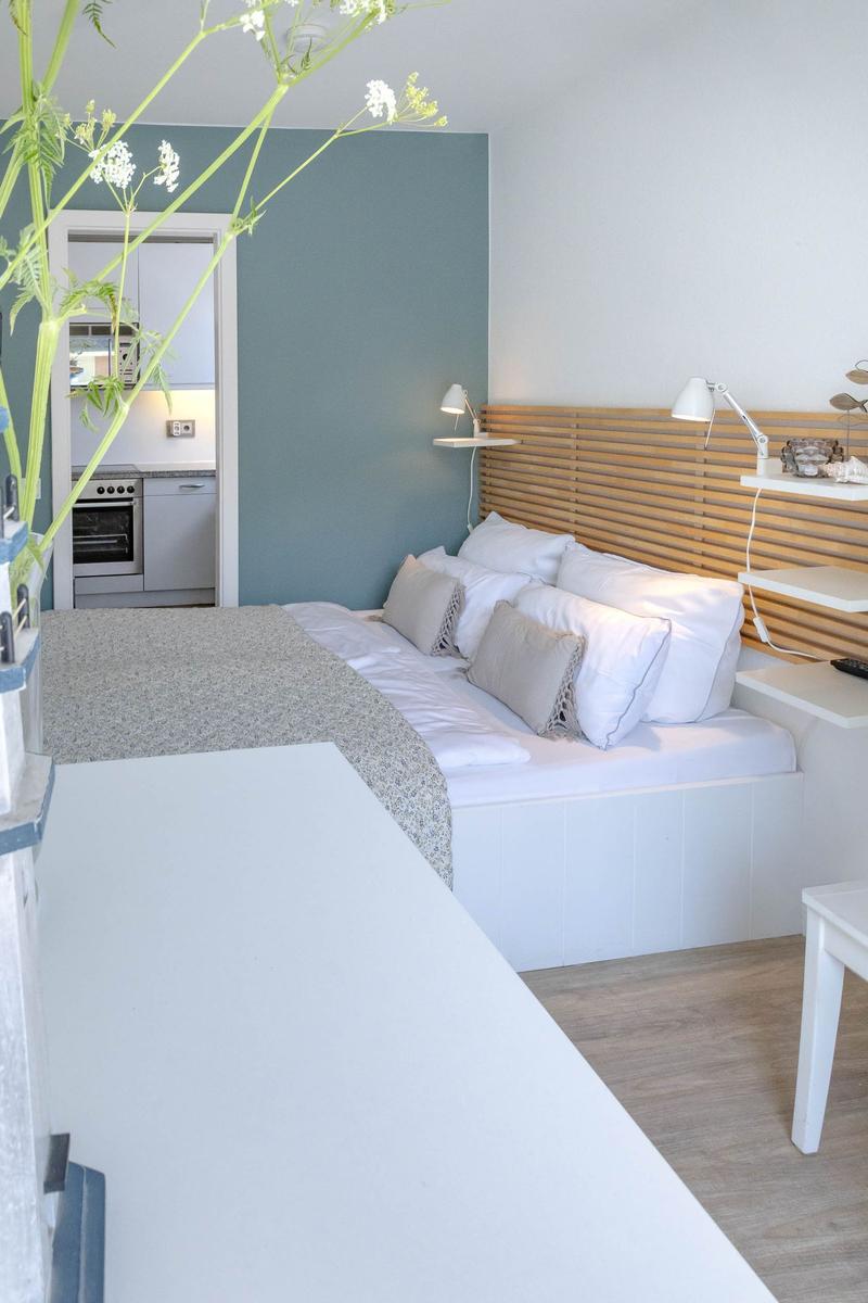Schlafzimmer mit Bett, Holz-Kopfteil und Nachttisch. Küche im Hintergrund sichtbar.