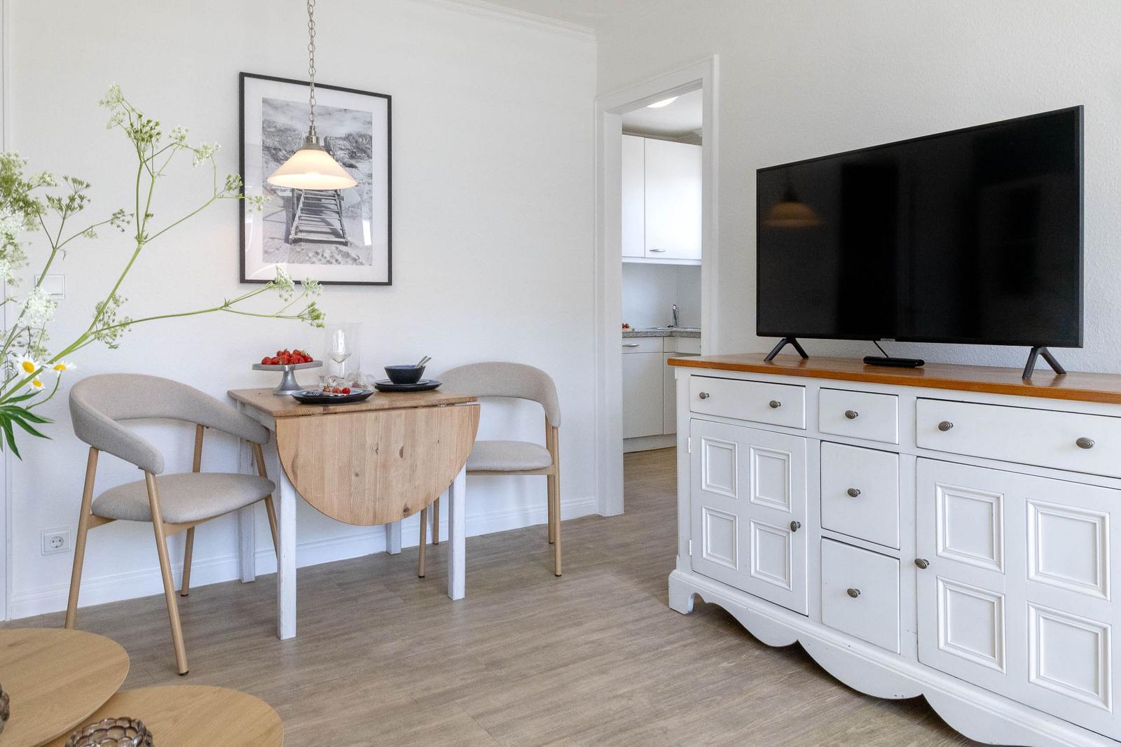 Essbereich mit Tisch, zwei Stühlen und Fernseher auf Schrank. Küche im Hintergrund sichtbar.