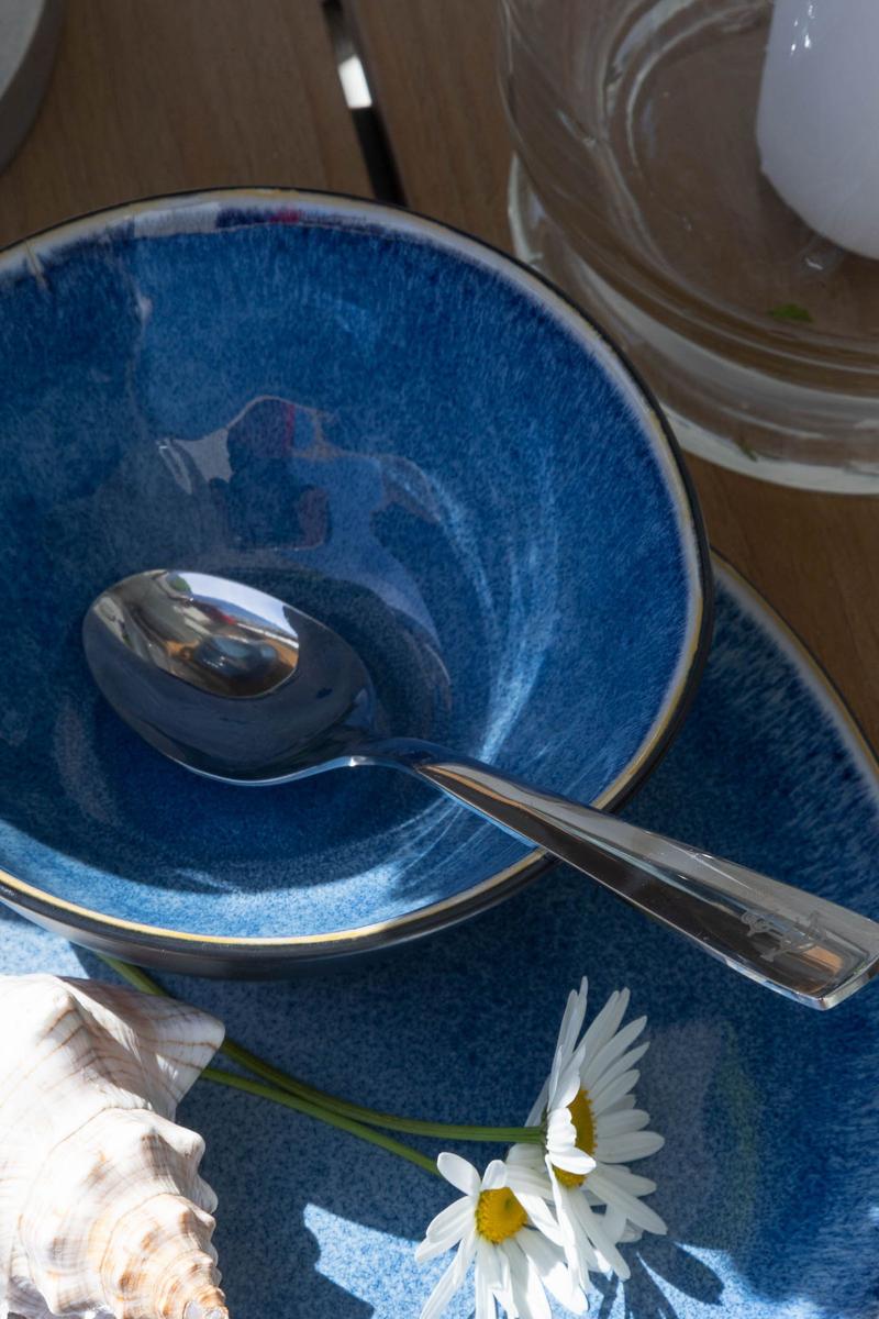 Blauer Teller mit Löffel, Kamille und Muschel auf Holztisch.