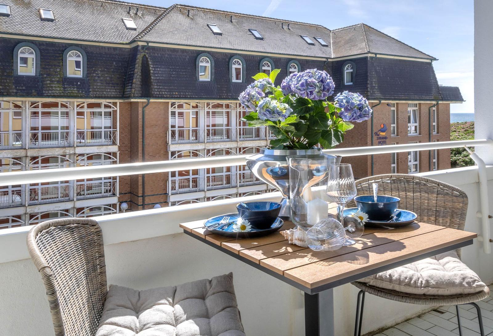 Balkon mit Tisch, Stühlen und Blumen. Blick auf Gebäude und Meer.