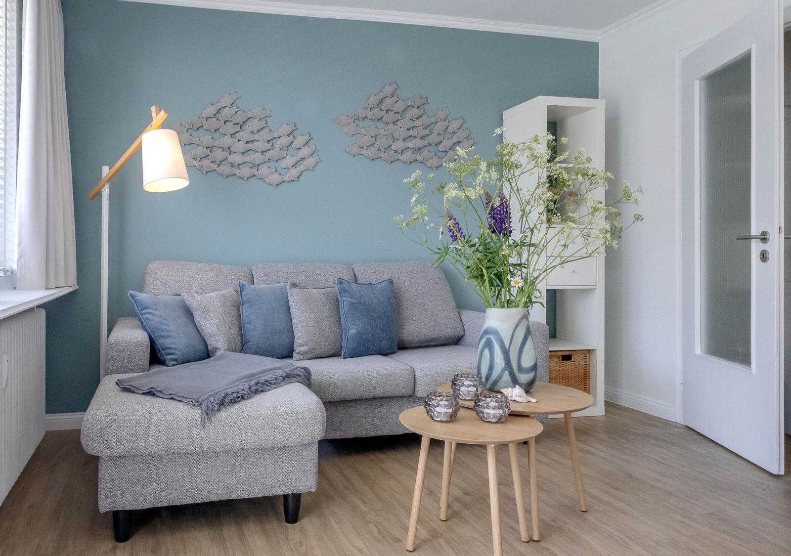 Ein Wohnzimmer mit grauem Sofa, blauer Wand und Holztisch. Blumen und Dekorationen sind sichtbar.