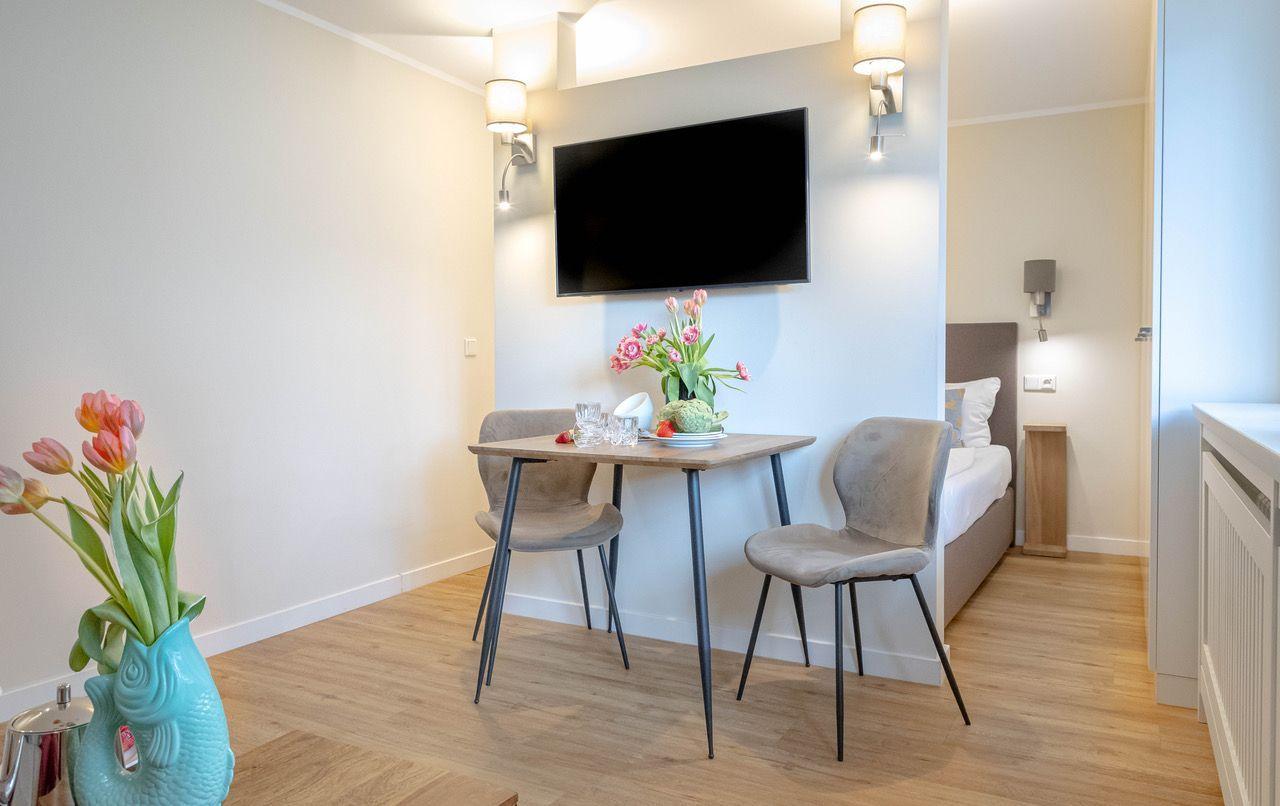 Ein Raum mit Tisch, zwei Stühlen, Fernseher und Bett im Hintergrund. Blumen und Holzboden.