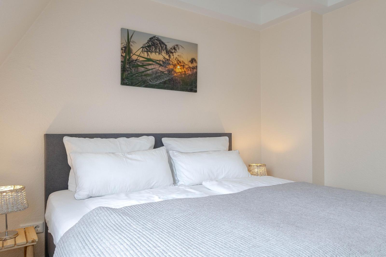Schlafzimmer mit Bett, Kissen und Wandbild. Beleuchtung durch Stehlampen.