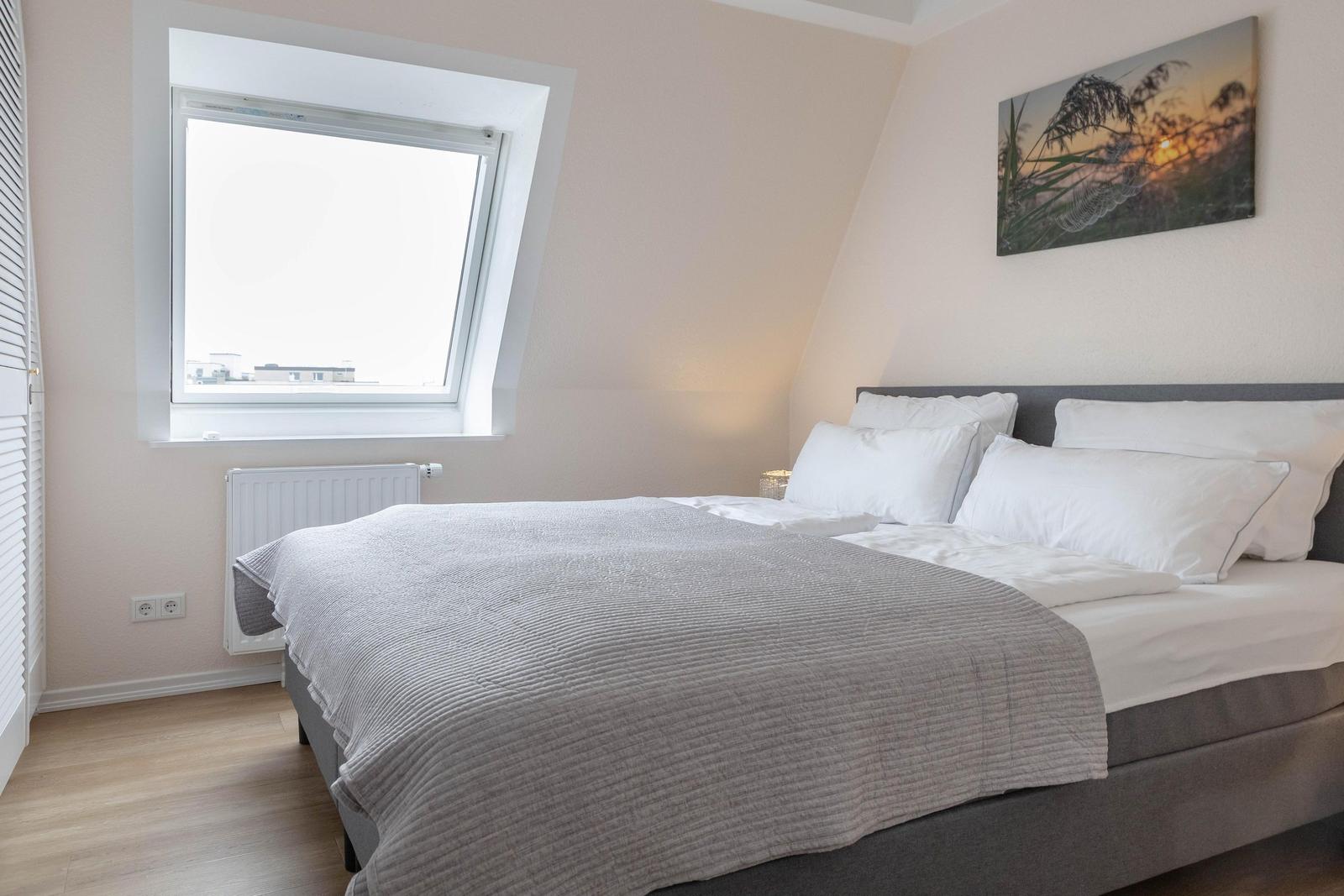Schlafzimmer mit Dachschrägen, Fenster und Bett mit grauer Bettdecke.