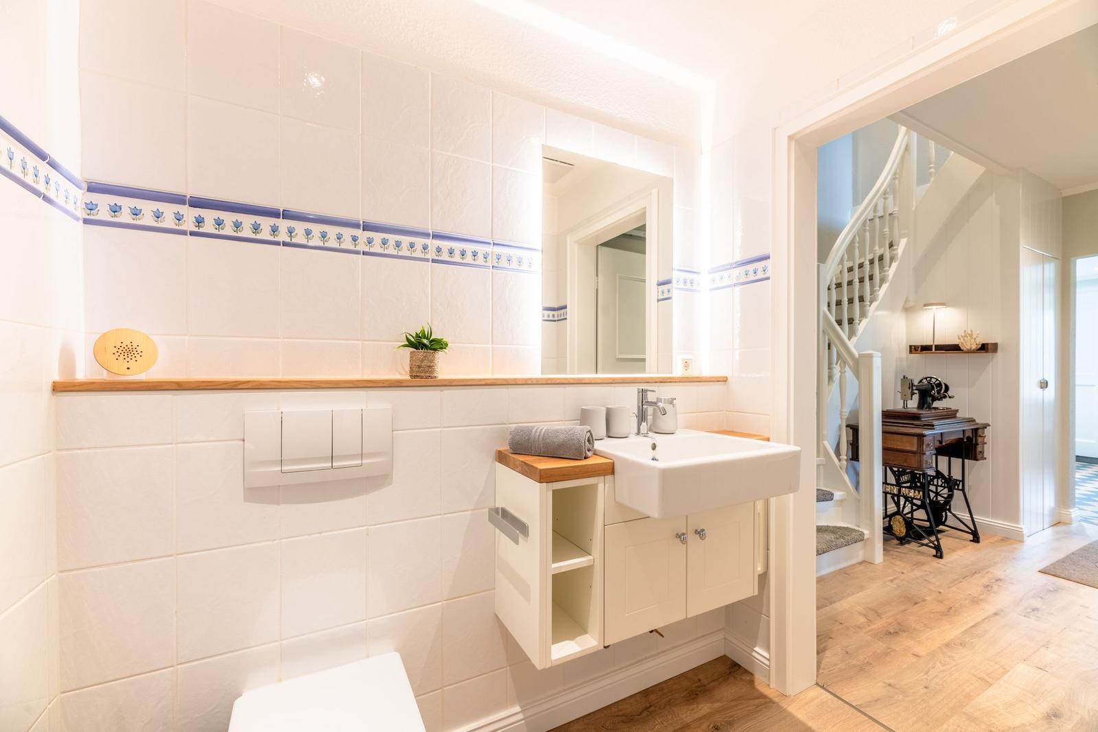 Badezimmer mit Waschbecken, Spiegel und Toilette. Hinterher ist eine Treppe zu sehen.