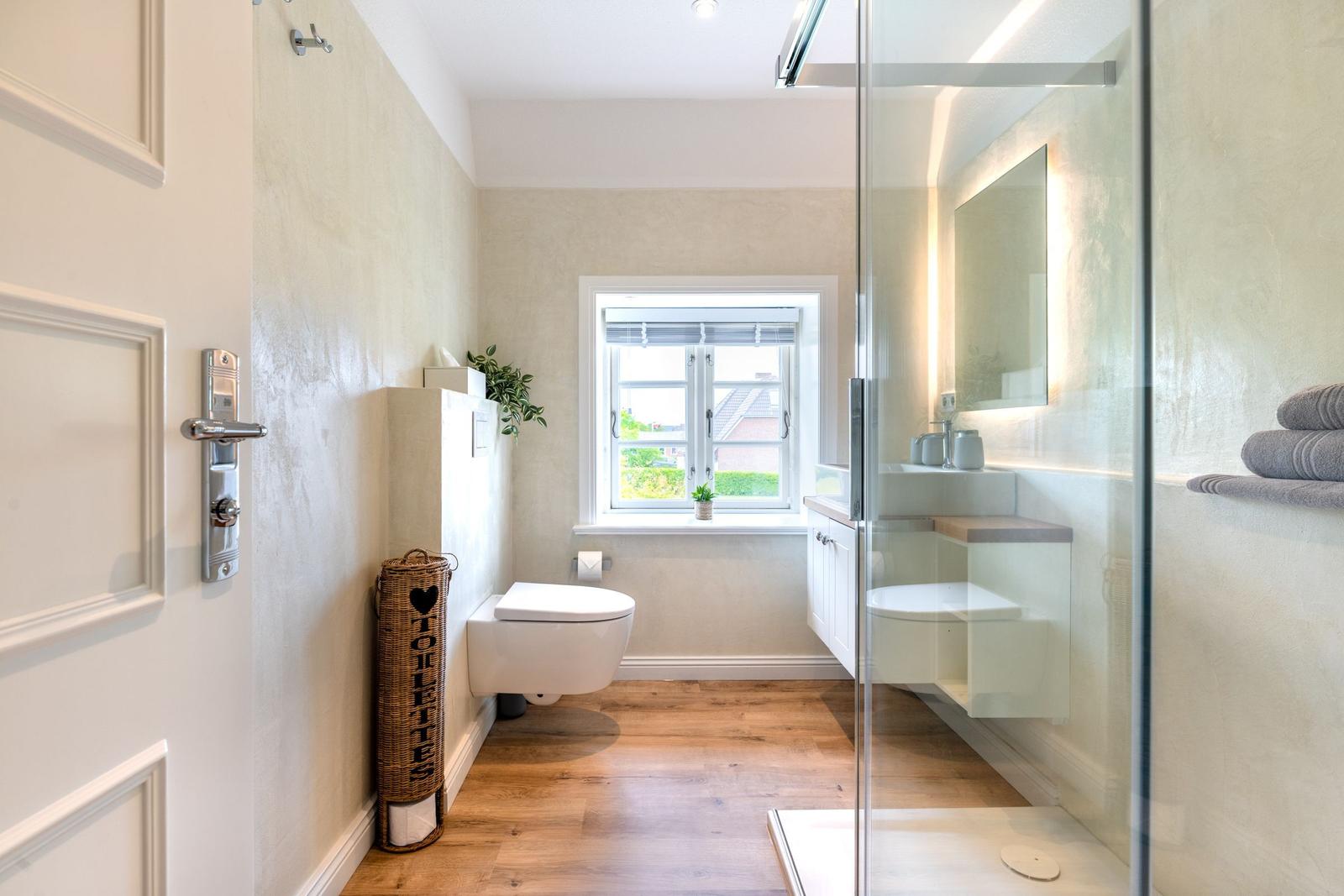Badezimmer mit Dusche, Toilette und Fenster. Holzboden und weiße Wände.