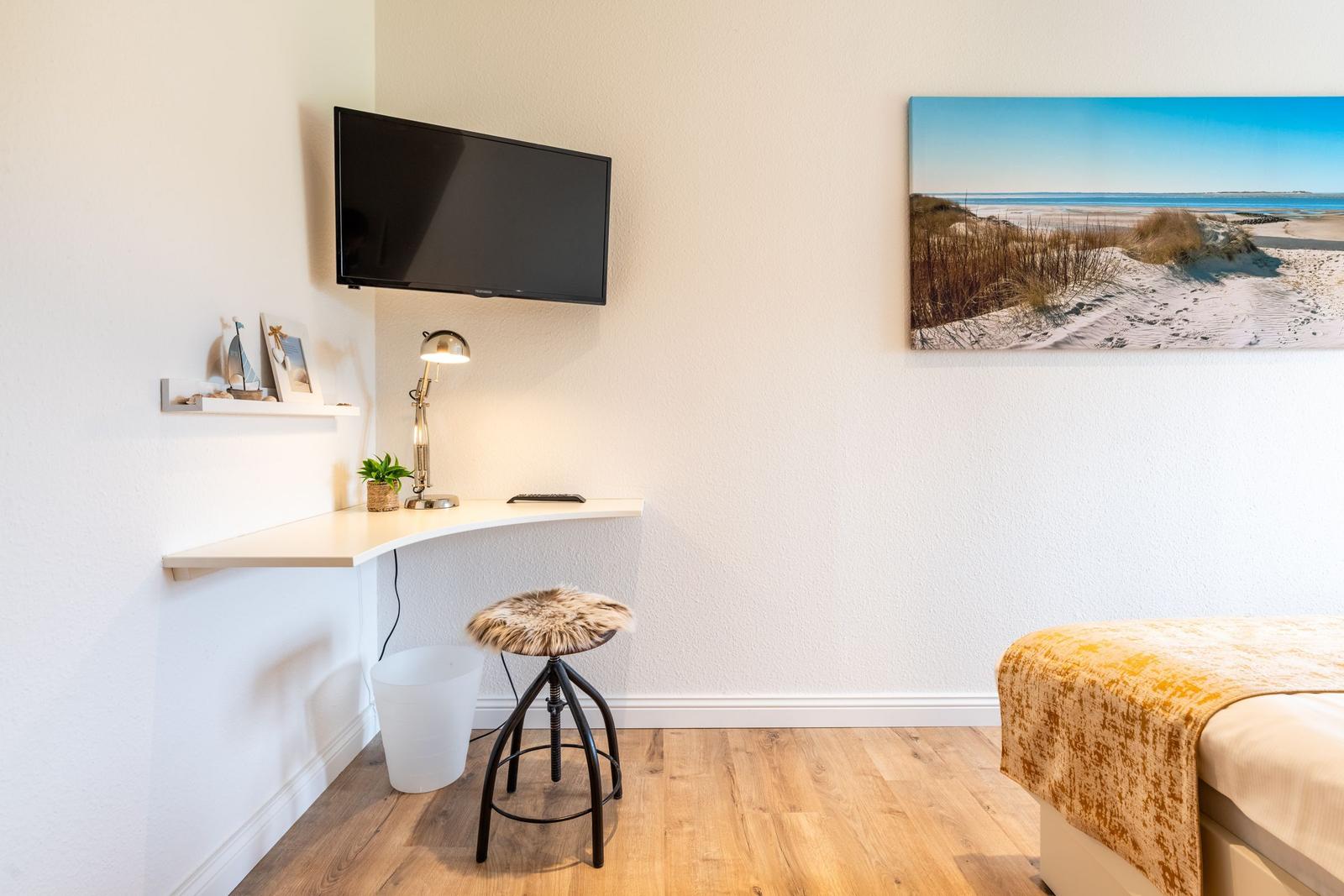 Schreibtisch mit Stuhl, Fernseher und Strandbild an der Wand.