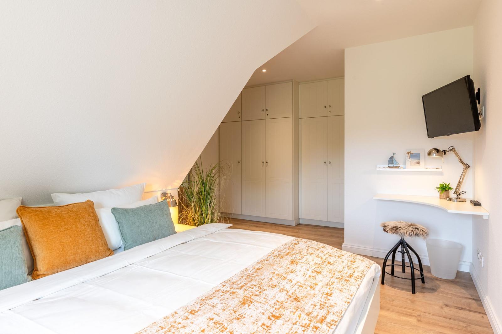 Schlafzimmer mit Dachschrägen, Bett, Kleiderschrank und Schreibtisch mit Fernseher.