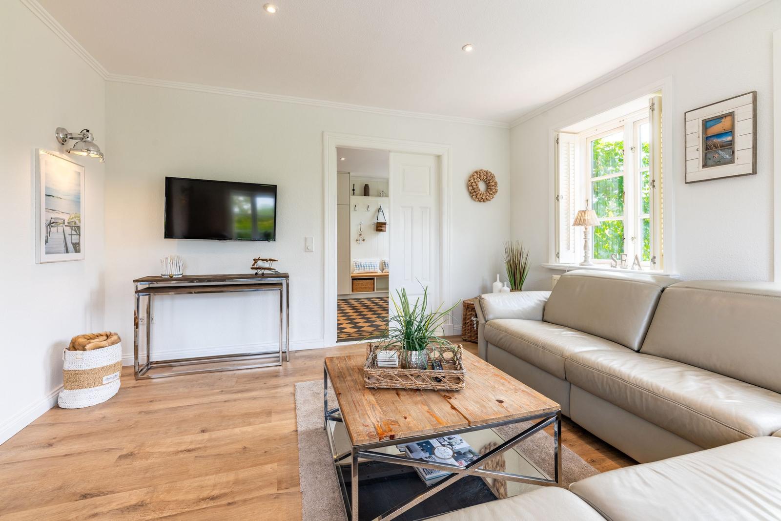 Wohnzimmer mit Sofa, Couchtisch und Fernseher. Fenster mit Blick auf Grün. Holzboden und Wandbilder.