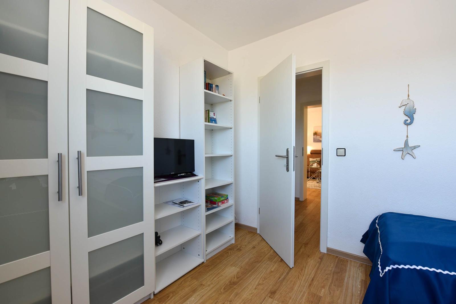 Schlafzimmer mit Bett, Kleiderschrank und Bücherregal. Tür führt in Wohnbereich.