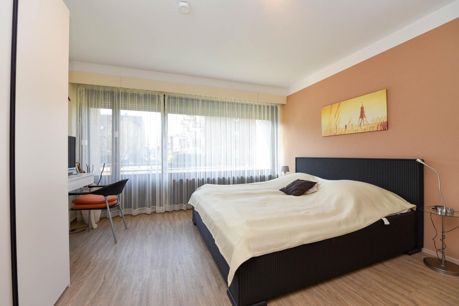 Schlafzimmer mit Bett, Schreibtisch und großem Fenster mit Blick auf die Stadt.