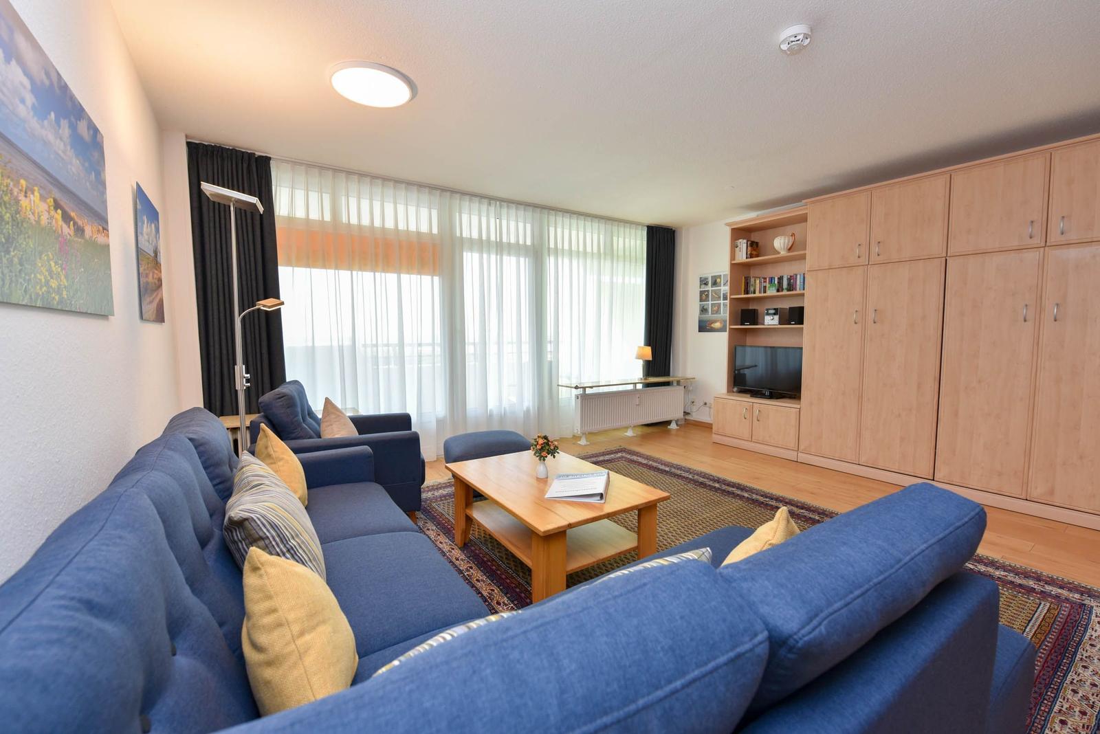 Wohnzimmer mit blauem Sofa, Holztisch und großen Fenstern.