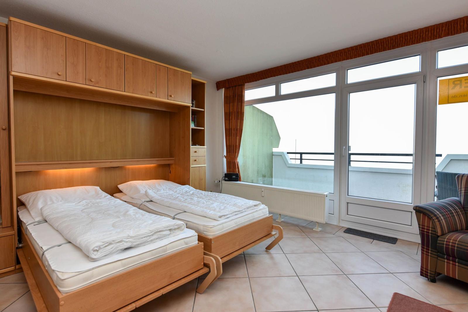 Zwei Schlafstellen mit Matratzen und weißen Bettwäschen. Holzmöbel, großes Fenster und Balkontür.