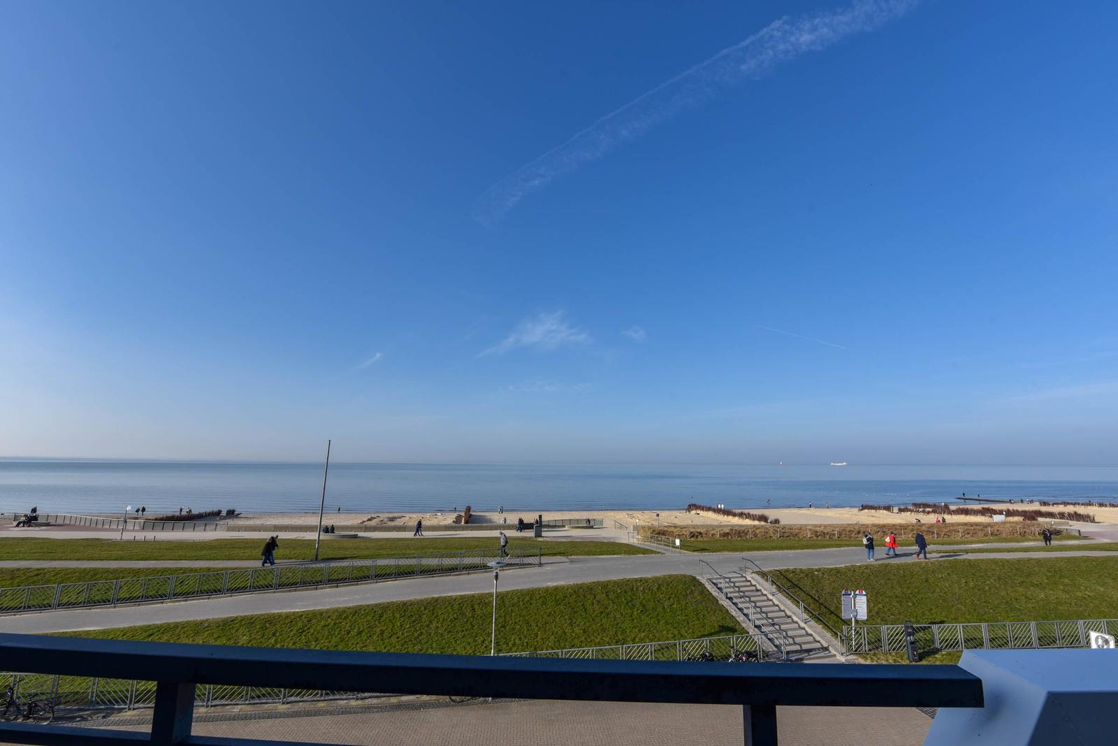 Blick auf Strand und Meer mit Grünflächen und Spaziergängern unter blauem Himmel.